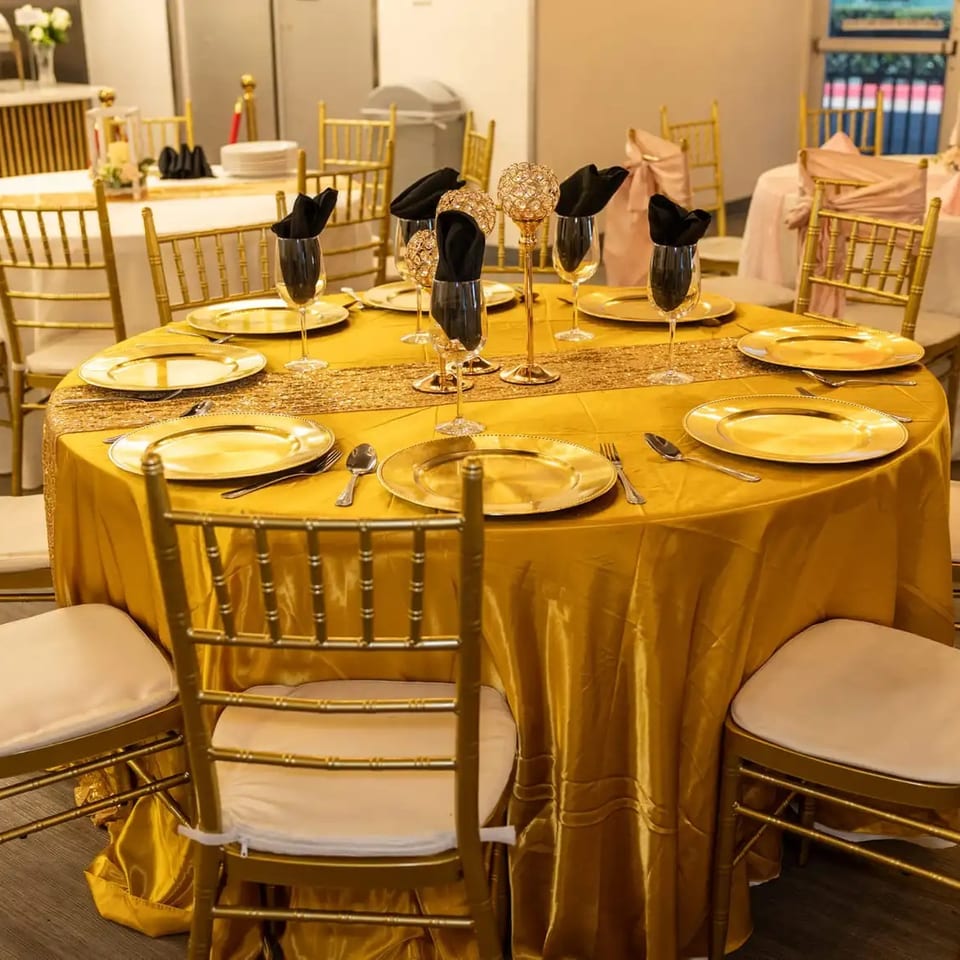 A round table set with golden plates, black napkins, and gold chairs in a banquet hall.