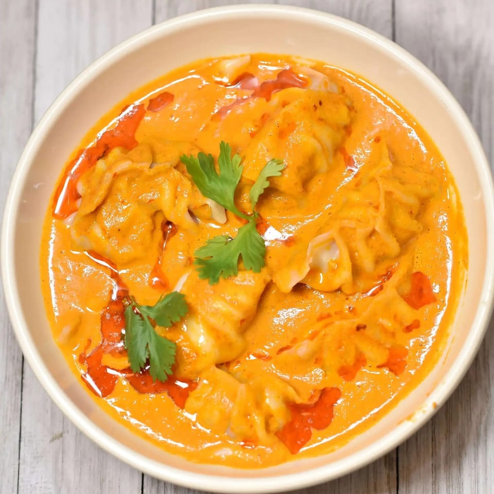 A bowl of momos in a vibrant orange sauce, garnished with fresh green cilantro.