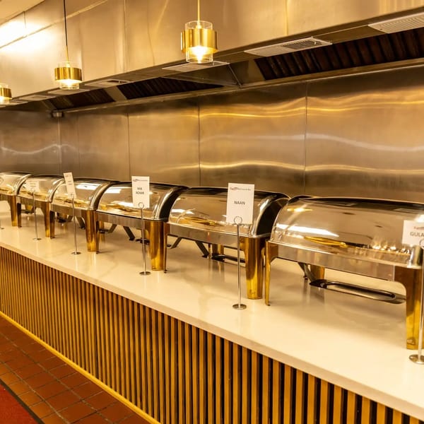 A buffet line with several silver chafing dishes on a white counter, with gold accents and industrial kitchen background.