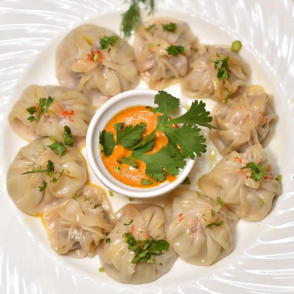 A white plate holds eight steamed momos surrounding a small bowl of orange dipping sauce garnished with cilantro.