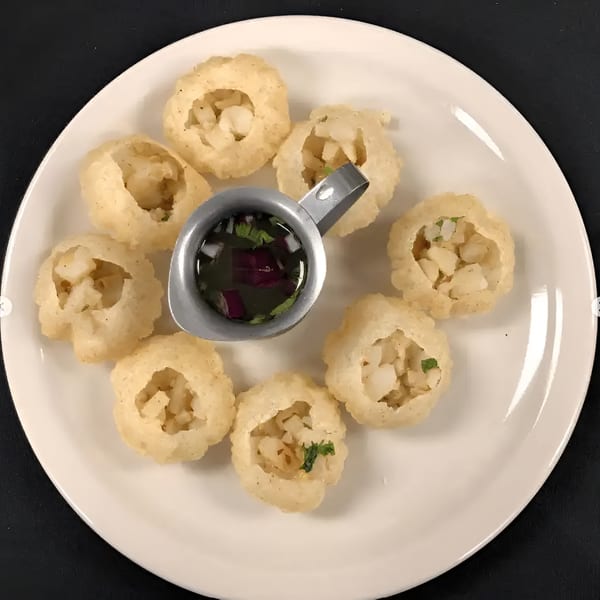 A white plate holds eight pani puri shells filled with potatoes, alongside a small pitcher of spiced water.
